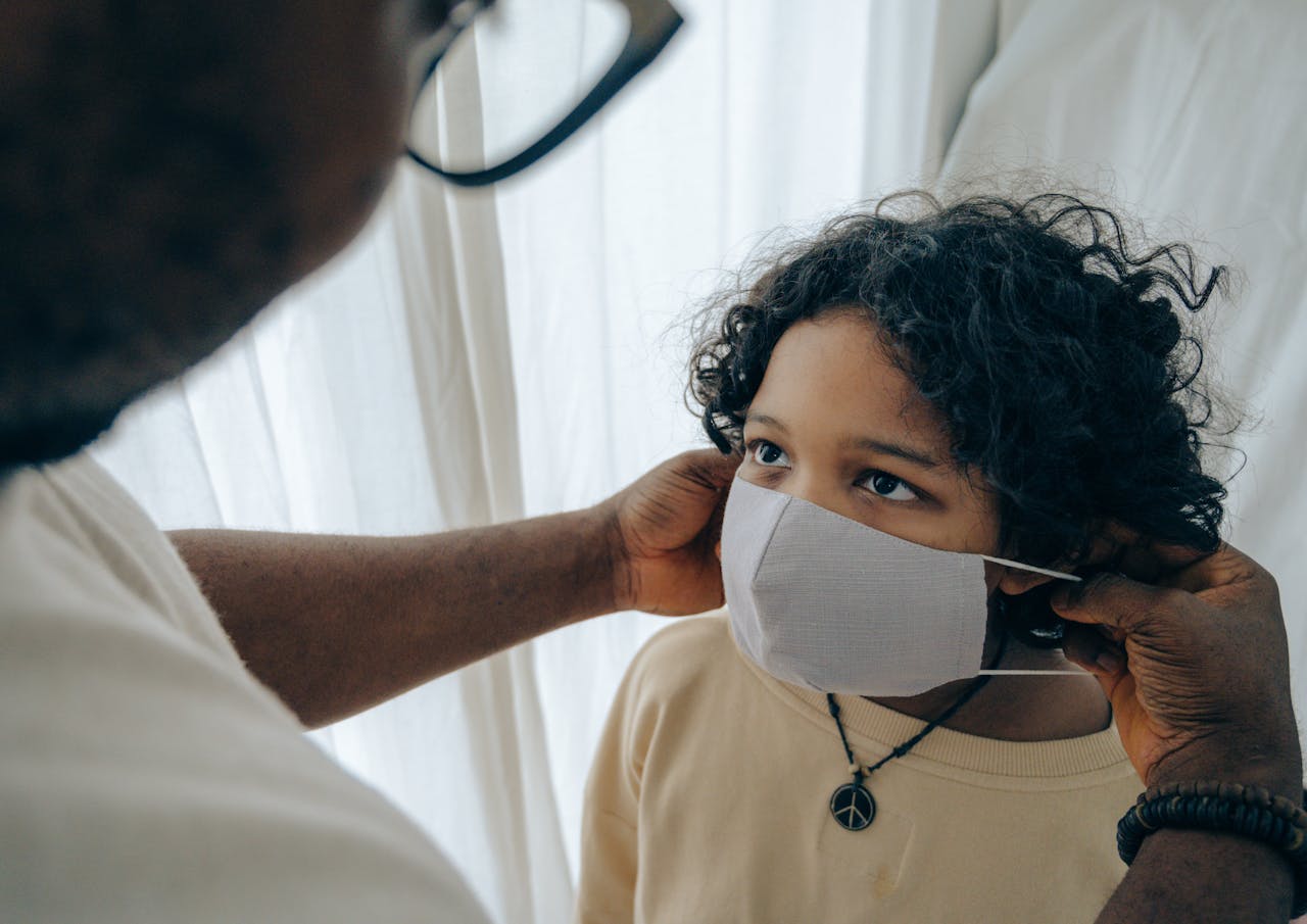 From above unrecognizable black man wearing protective mask on face of ethnic kid standing in room near window closed with curtain